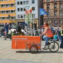 Woman riding a bike with a trailer labeled Reallabore through downtown.