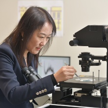Prof. Laura Na Liu stands at a microscope looks into a microscope.