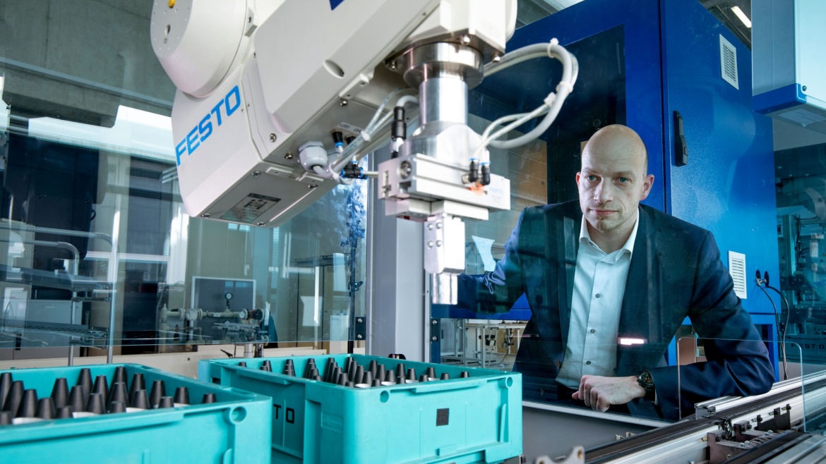 Prof. Huber stands besides an assembly line. He is looking at a robot-arm in movement.