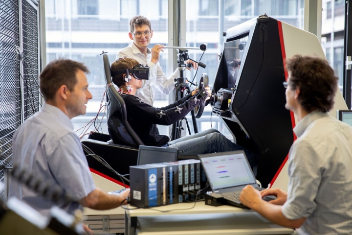 A man sits in the driving simulator. Prof. Jörg Fehr is standing behind him, with two colleagues in the foreground.