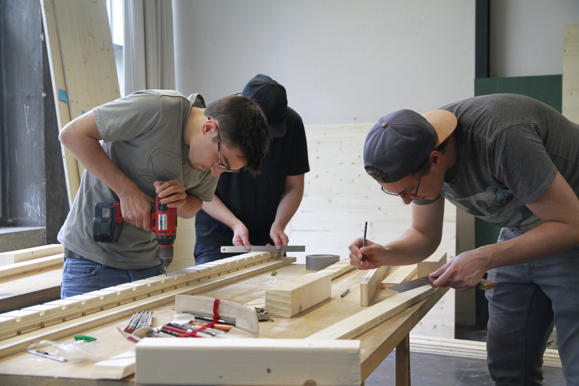 An einem Arbeitstisch werken drei Studenten mit Holz. Einer hält eine Bohrmaschine in der Hand, einer ein Lineal und einer einen Bleistift.