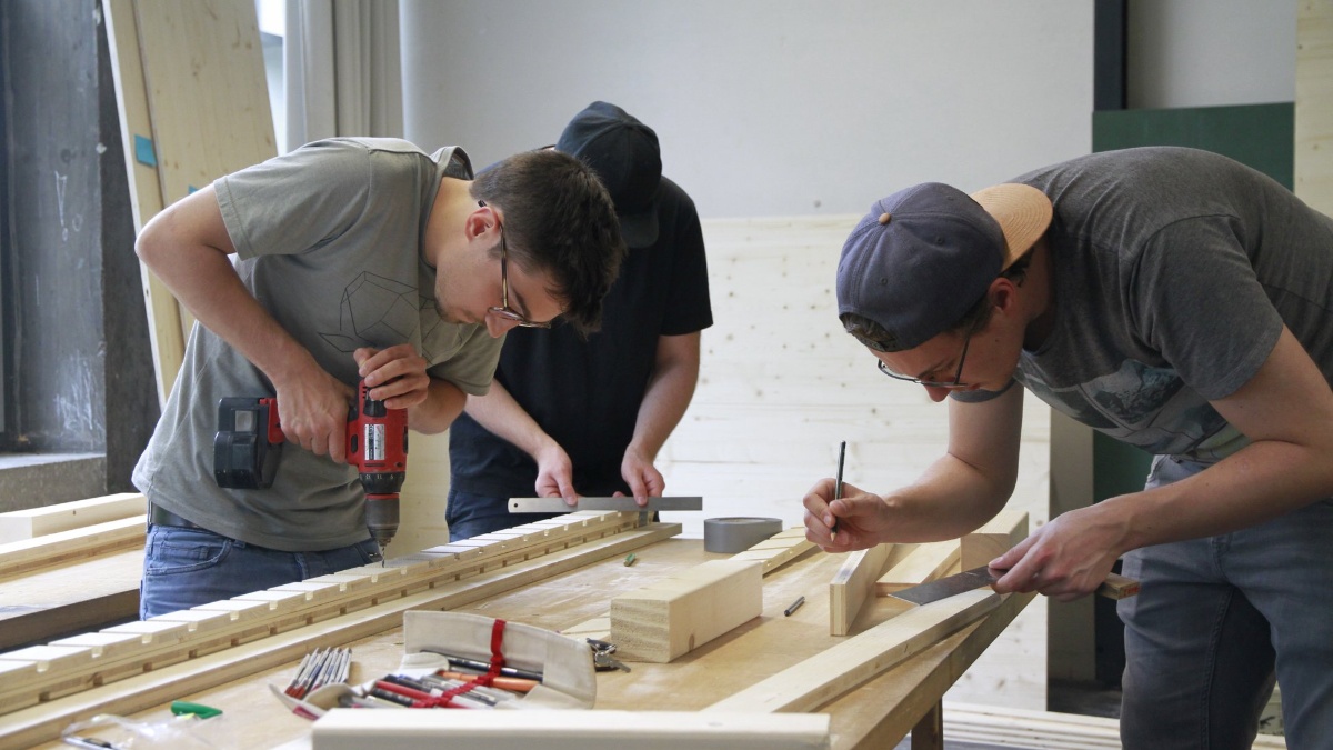 An einem Arbeitstisch werken drei Studenten mit Holz. Einer hält eine Bohrmaschine in der Hand, einer ein Lineal und einer einen Bleistift.