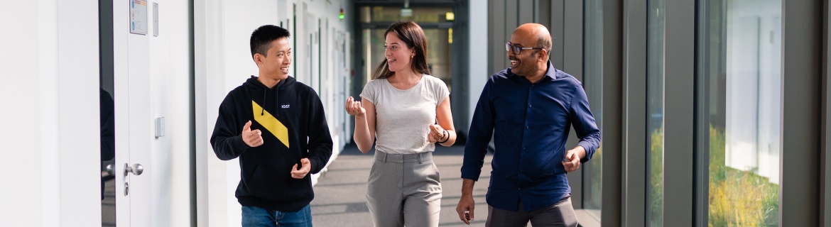 Drei Studierende gehen gemeinsam durch einen modernen Hochschulflur mit großen Fensterfronten. Sie unterhalten sich angeregt und wirken gut gelaunt. Die Szene vermittelt eine offene und kollaborative Lernatmosphäre.“