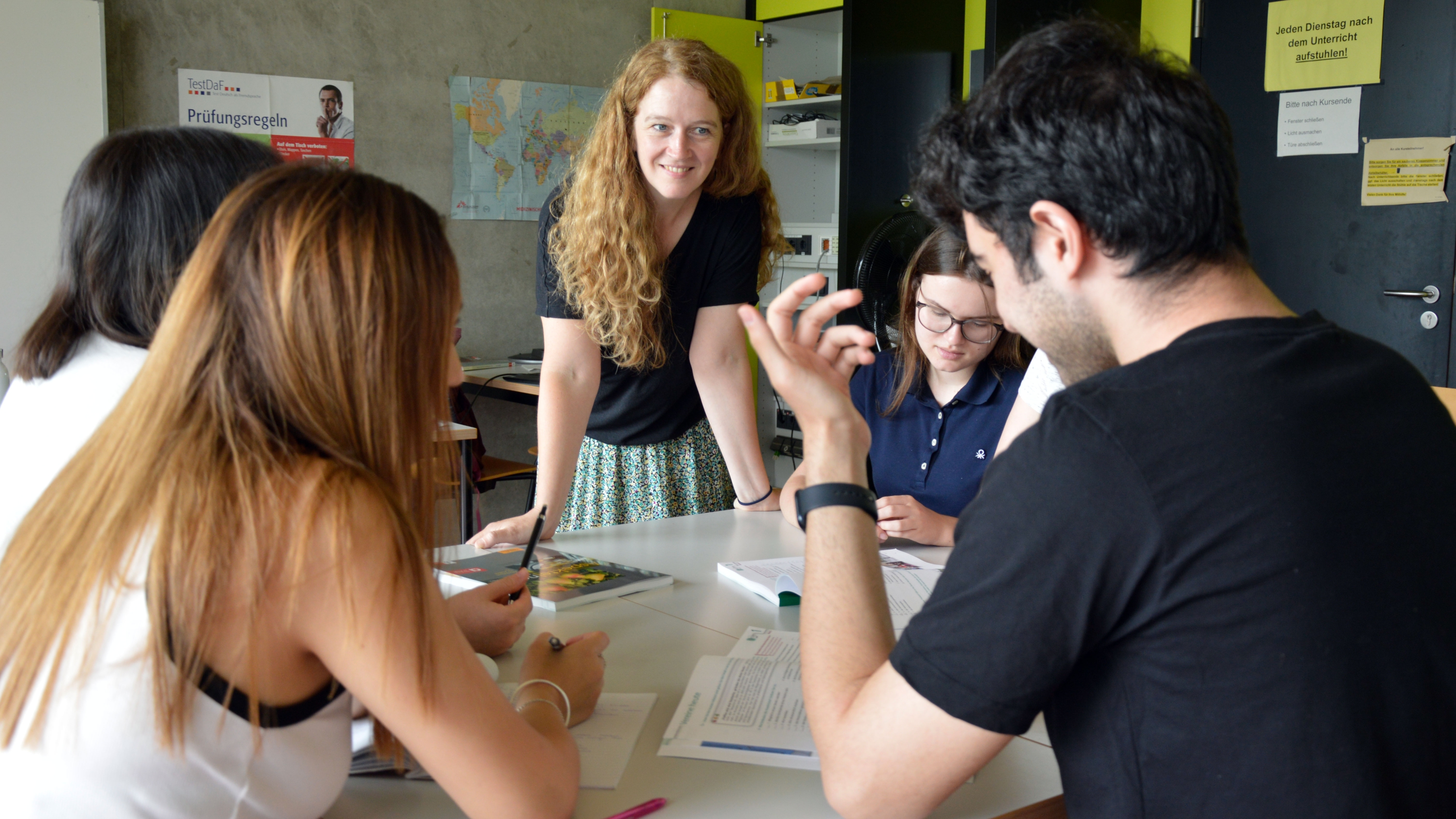 Students are sitting at a table. A lecturer stands opposite them and listens attentively to the students.