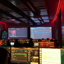 Ein Kontrollraum zur Überwachung von Satelliten. Zwei Personen sitzen mit dem Rücken zur Kamera gewandt vor Bildschirmen. Der Raum ist abgedunkelt und liegt in rotem Licht.