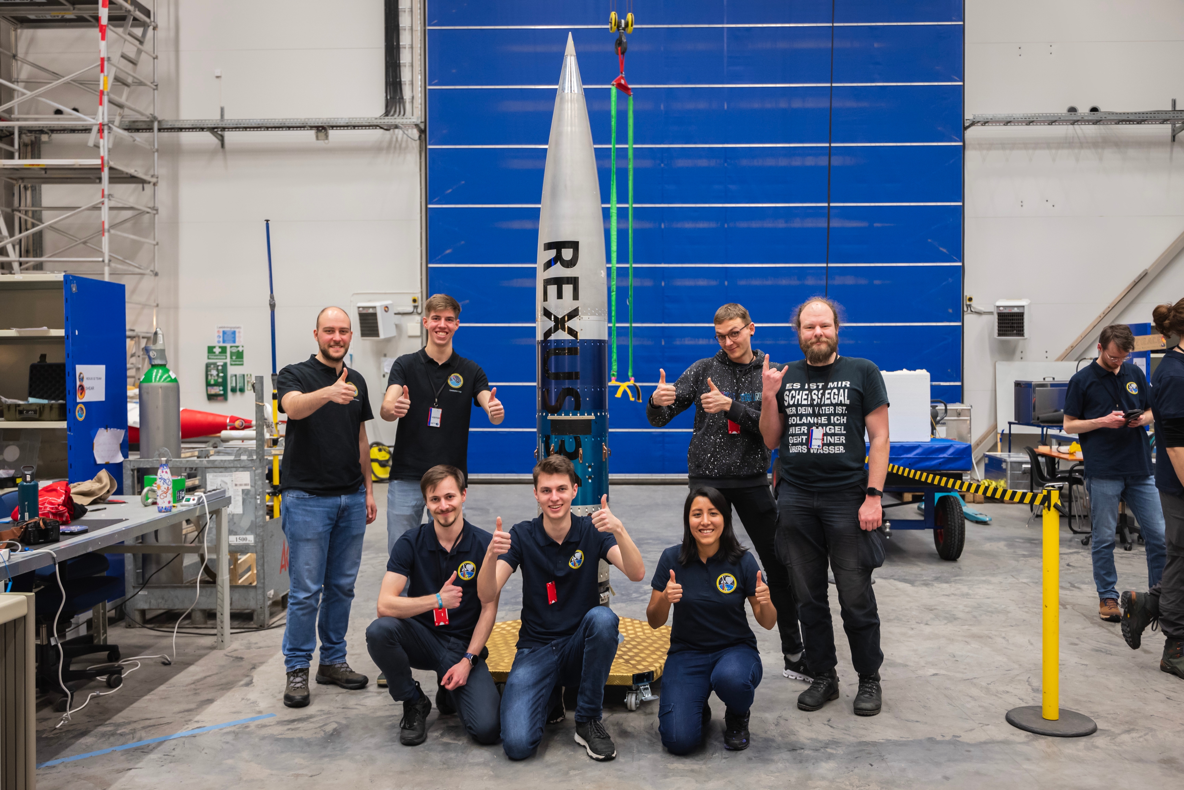 A group of students posing for a photo infront of the REXUS rocket in a warehouse.