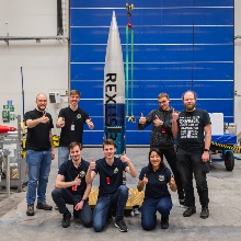 A group of students posing for a photo infront of the REXUS rocket in a warehouse.