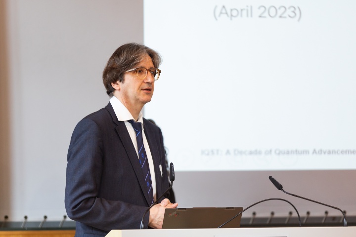 Professor Fedor Jelezko from IQST speaks at the podium on stage. A PowerPoint presentation can be seen in the background. 