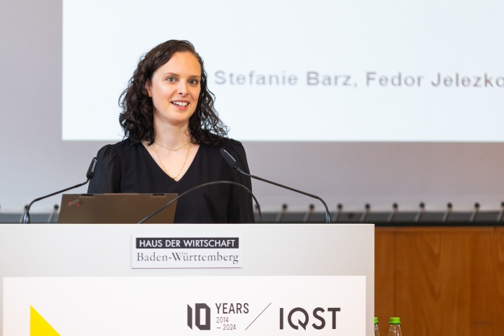 Professorin Stefanie Barz von IQST spricht am Rednerpult auf der Bühne. Im Hintergrund ist eine PowerPoint Präsentation zu sehen. 
