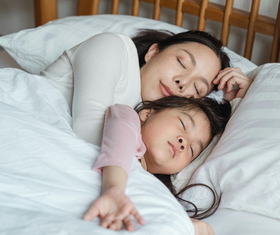 Eine Mutter und ihr Kind liegen gemeinsam schlafend im Bett, zugedeckt mit einer weißen Bettdecke.