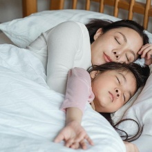 Eine Mutter und ihr Kind liegen gemeinsam schlafend im Bett, zugedeckt mit einer weißen Bettdecke.