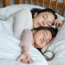 A mother and her child lie asleep together in bed, covered with a white comforter.
