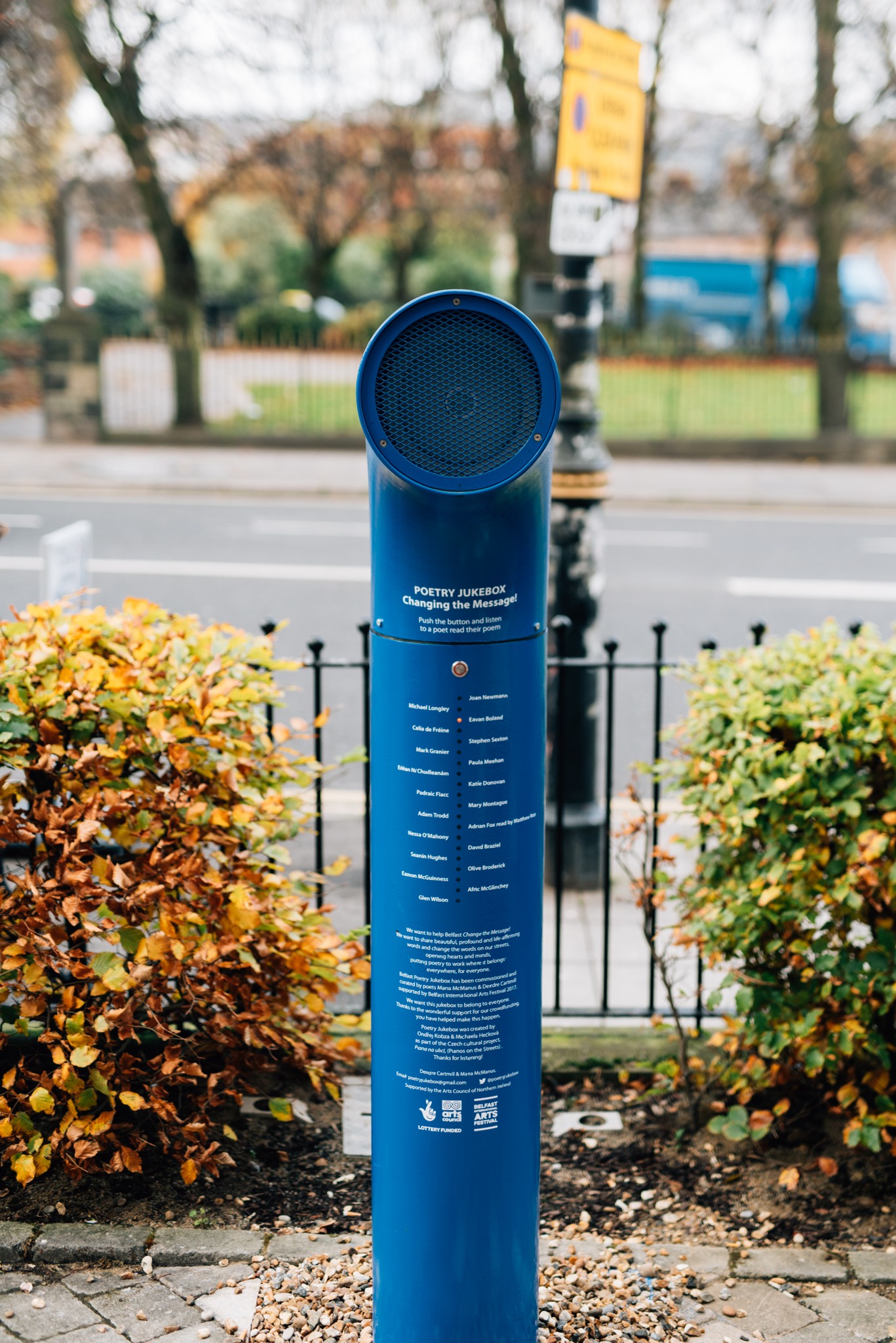 Das Bild zeigt die Jukebox, aufgestellt in einer Parkanlage, vor einem Zaun in Richtung Straße. Die Jukebox sieht aus, wie eine dicke blaue Säule, das aus dem Boden schießt, am oberen Ende mit einem 90-Grad-Bogen. Auf der Säule stehen in weißer Schrift die Namen der Dichter*innen und ihrer Gedichte.