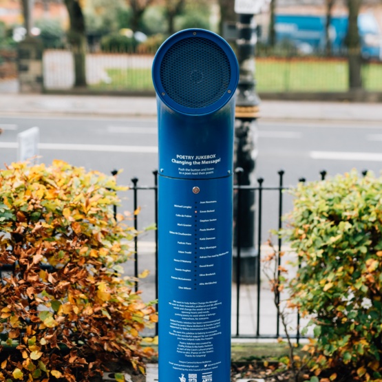 Das Bild zeigt die Jukebox, aufgestellt in einer Parkanlage, vor einem Zaun in Richtung Straße. Die Jukebox sieht aus, wie eine dicke blaue Säule, das aus dem Boden schießt, am oberen Ende mit einem 90-Grad-Bogen. Auf der Säule stehen in weißer Schrift die Namen der Dichter*innen und ihrer Gedichte.