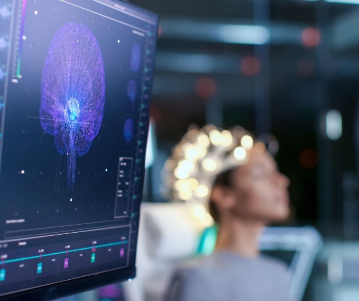 Im Hintergrund ist verschwommen eine Person zu sehen, die eine EEG-Kappe trägt. Im Vordergrund ein Computerbildschirm, auf dem eine Animation eines Gehirns zu sehen ist.