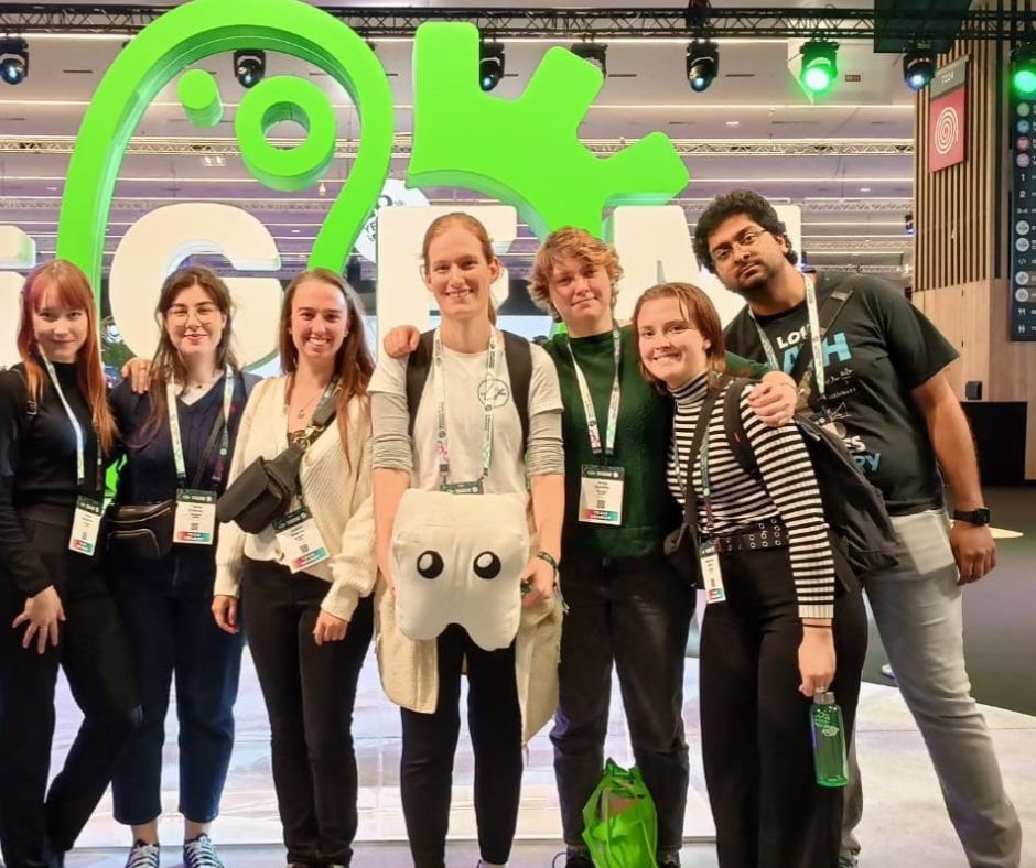 Das Team posiert vor einem grünen iGEM Aufsteller gemeinsam mit dem Teammaskottchen "Zahni".