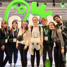 Das Team posiert vor einem grünen iGEM Aufsteller gemeinsam mit dem Teammaskottchen "Zahni".