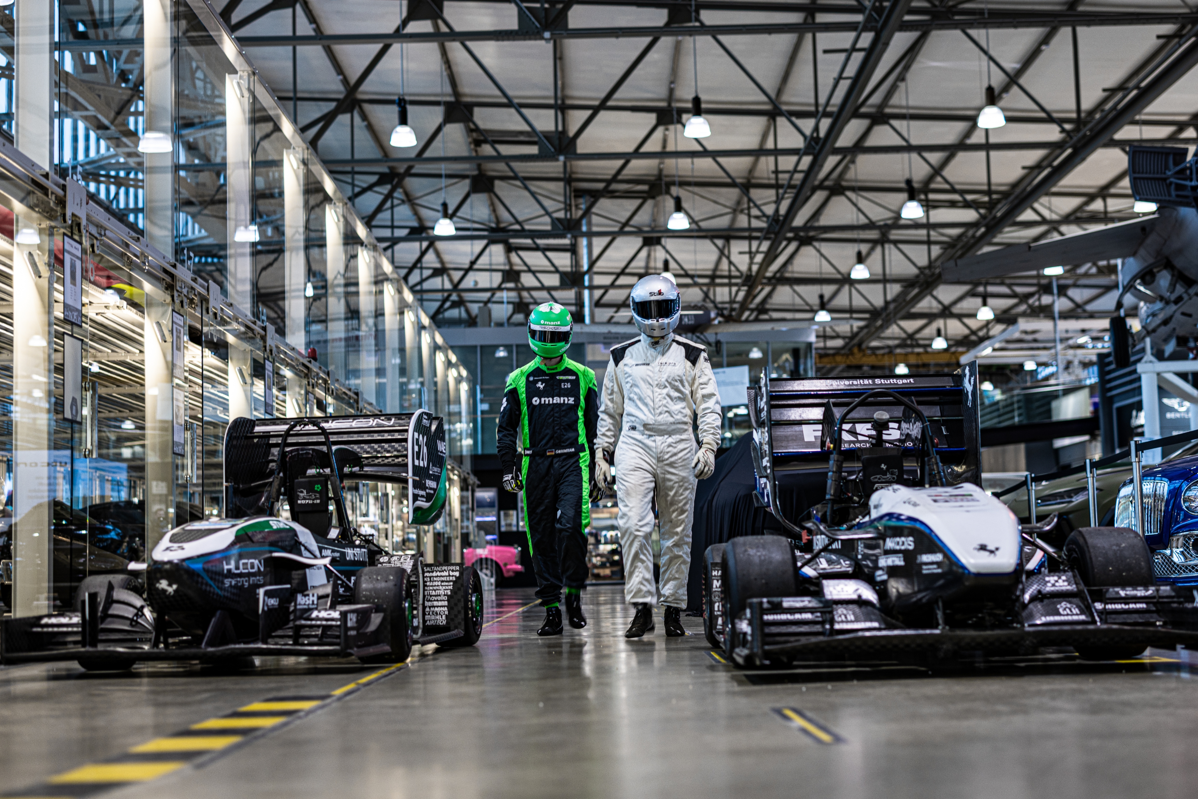 Zwei Rennfahrer gehen in Rennfahrerkluft und Helm zwischen zwei Rennfahrzeugen hindurch. Die Fahrzeuge stehen in einer Lagerhalle. 