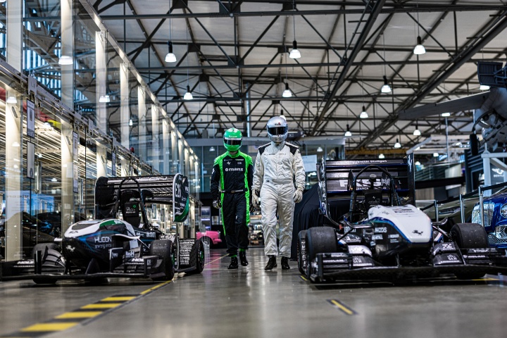 Two racing drivers in racing gear and helmets walk between two racing cars. The vehicles are parked in a warehouse.