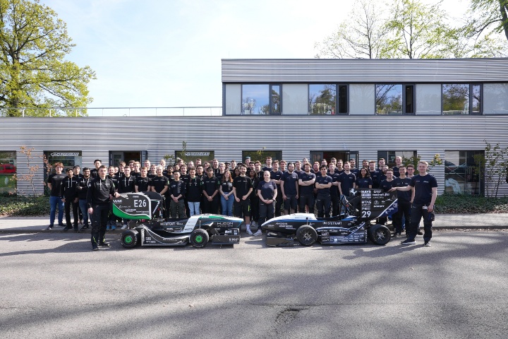 Die Studierenden posieren für ein Gruppenfoto vor der Basis des Rennteams auf dem Campus Vaihingen sowie zwei Rennfahrzeugen.