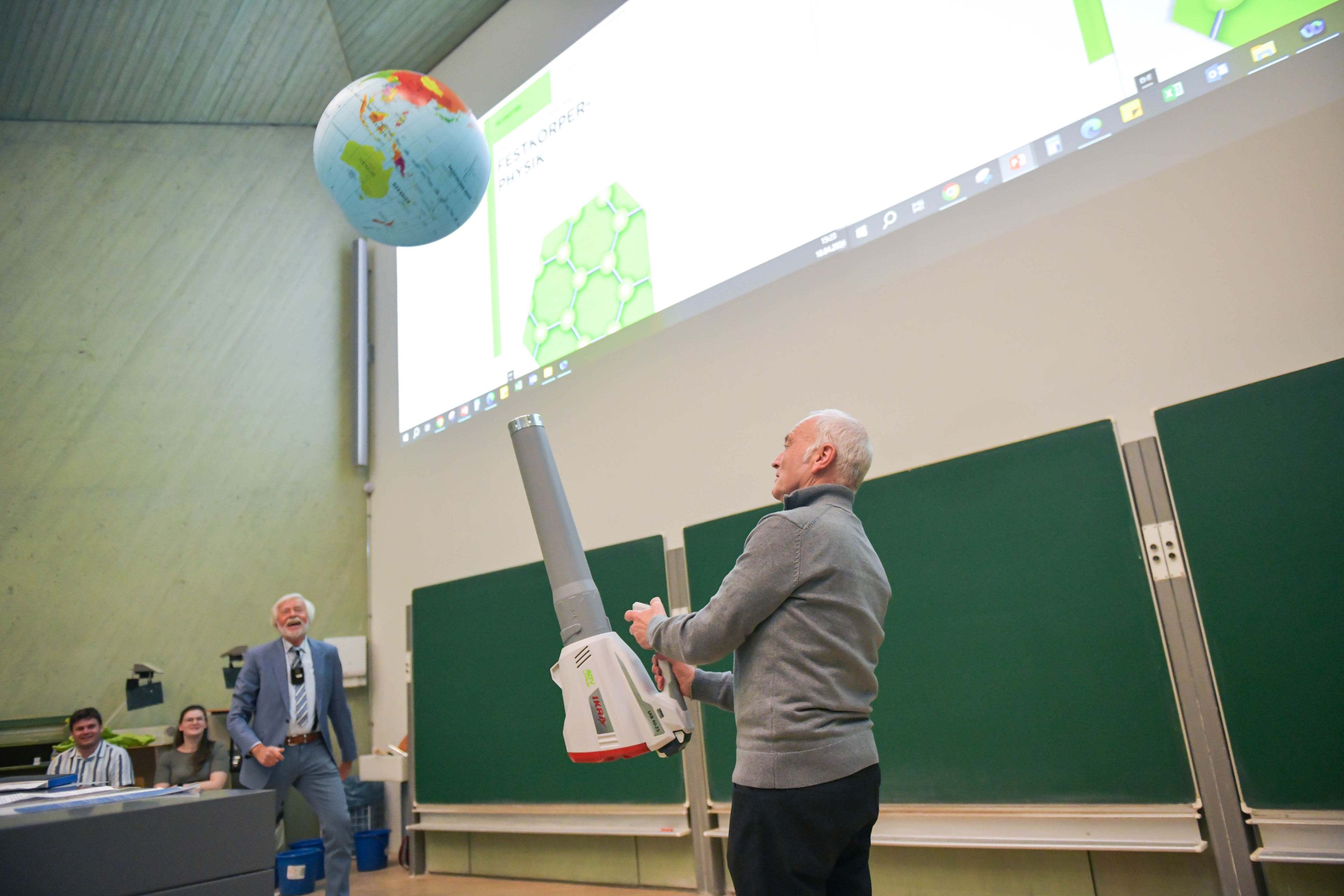 Ulrich Schneider steht in einem großen Hörsaal und hält mit einem Blasgerät einen Ball in Gestalt einer Weltkugel in der Luft.