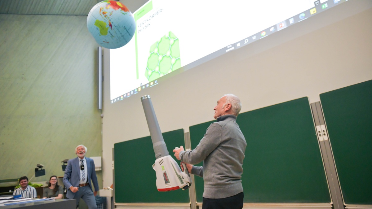Ulrich Schneider steht in einem großen Hörsaal und hält mit einem Blasgerät einen Ball in Gestalt einer Weltkugel in der Luft.