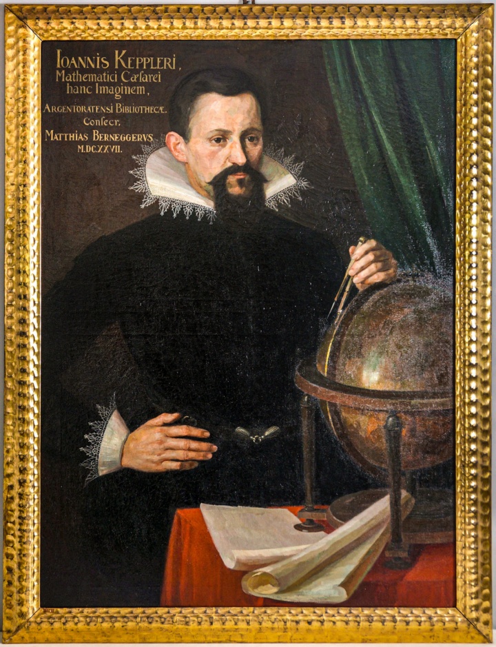Johannes Kepler ein Mann mit dunklem Bart, Talar und Renaissancekragen steht an einem Tisch mit roter Decke. Seine Linke Hand berührt einen bronzenem Himmelsglobus. Seine rechte Hand stützt er in die Seite. Auf dem Tisch liegen alte Bücher und Schriften.