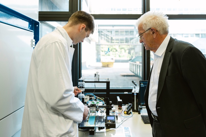 A student employee (right) in a white coat shows the Minister President (right) his laboratory workstation.