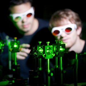 Two men wearing google with red shining lenses are looking into a green laser produced by a laser machine.