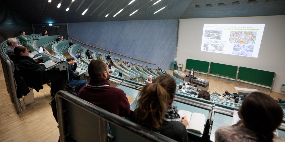 Ein Hörsaal voller Menschen, die einen Vortrag verfolgen. Der Vortragende, Professor Frank Allgöwer, ist im Hintergrund des Bildes zu sehen.