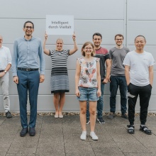 Eight people of different age, size, gender and origin. In the middle, a sign is held up with the inscription "Diversity promotes intelligence".". 