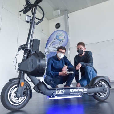 Zwei Wissenschaftler knien auf dem Boden und blicken auf einen im Bildvordergrund abgebildeten E-Scooter