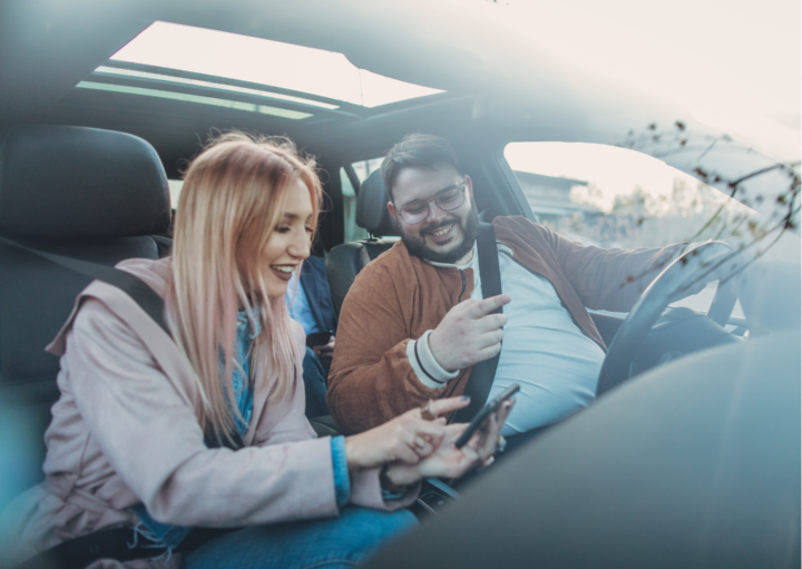 Zwei Personen sitzen in einem Auto. Fahrer und Beifahrerin schauen sich gemeinsam etwas auf dem Smartphone an. 
