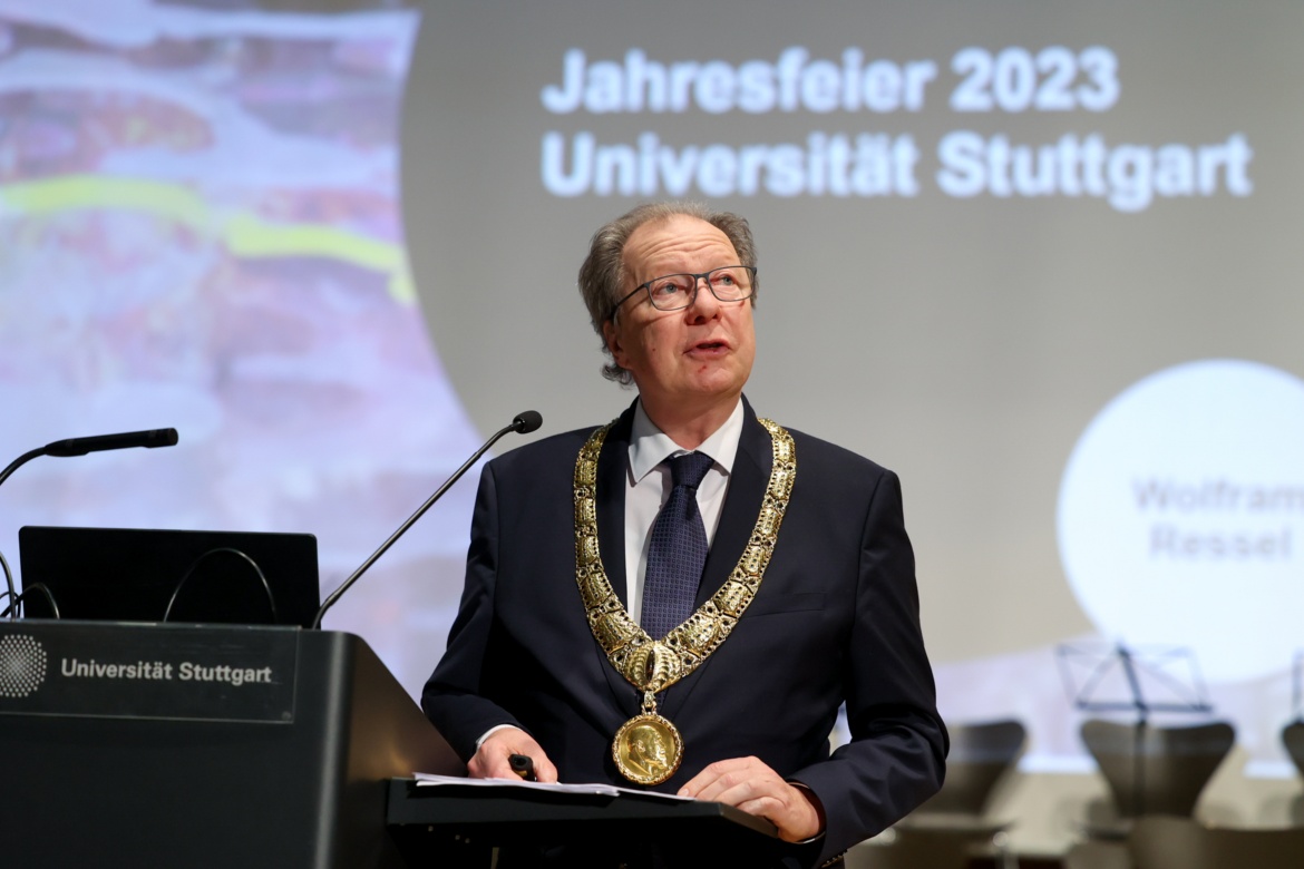 Rektor Wolfram Ressel am Rednerpult, er trägt einen Anzug und hat einen großen goldenen Orden um den Hals. Hinter ihm ist mit Beamer ein Bild an die Wand geworfen, das die Aufschrift "Jahresfeier 2023 Universität Stuttgart" trägt.