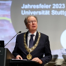 Rektor Wolfram Ressel am Rednerpult, er trägt einen Anzug und hat einen großen goldenen Orden um den Hals. Hinter ihm ist mit Beamer ein Bild an die Wand geworfen, das die Aufschrift "Jahresfeier 2023 Universität Stuttgart" trägt.