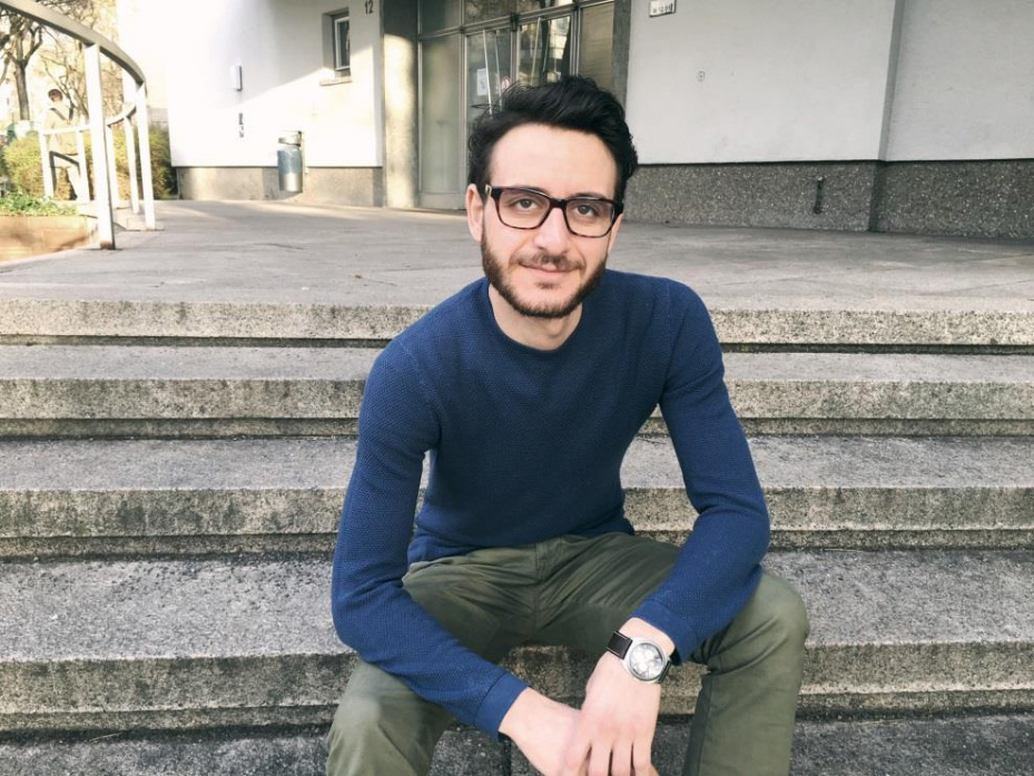 Oualid Ben Rejeb ist ein junger Mann mit dunklen Haaren, kurzem Vollbart und einer kastigen Brille. Er sitzt lässig auf einer Treppenstufe und blickt entspannt in Richtung seines Gegenübers.