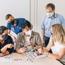 A five-member working group of different genders and with and without a migration background gathers around a table and discusses with each other. In the middle sits a young man holding a model of a molecule in his hand.