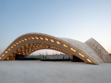 Arched, open wooden pavilion with segmented hollow cassette structure on the grounds of the Federal Horticultural Show in Heilbronn 2019, where the pavilion served as an event space with a multipurpose stage. 
