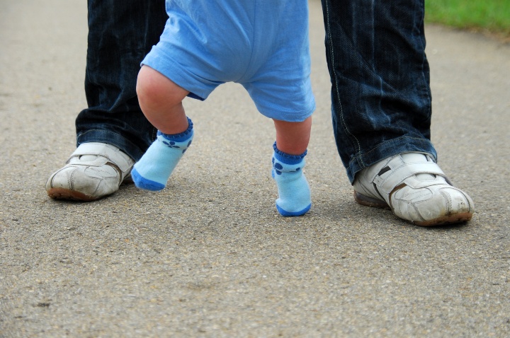 Symbolbild: Ein Kleinkind in blauen Socken lernt laufen, indem es von einem Elternteil geführt wird.